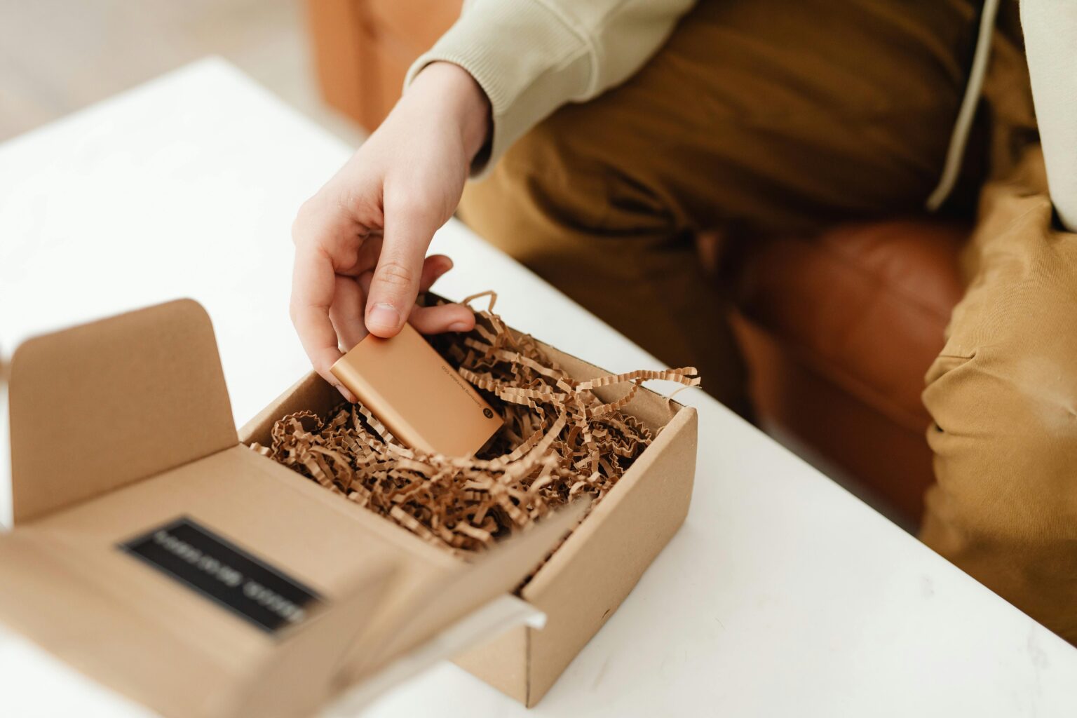 A person unboxes a small item from a cardboard package filled with crinkle paper, indoors.